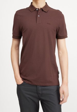 Homme portant un polo marron à manches courtes avec deux boutons et un pantalon noir, une main dans la poche.