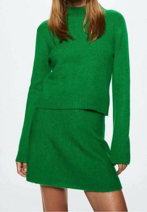 Femme portant un pull en maille vert à manches longues et une mini-jupe assortie en maille verte, sur un fond uni.