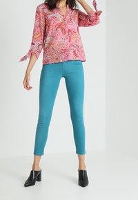 Růžová blůza s paisley vzorem a véčkovým výstřihem, se zavazovacími rukávy, zkombinovaná s úzkými tyrkysovými kalhotami a černými koženými mules na podpatku.