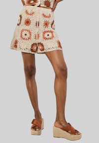 Pantaloni corti all'uncinetto con una base beige caratterizzati da motivi floreali in ruggine e crema. Abbinati a sandali con zeppa marroni con suola intrecciata.