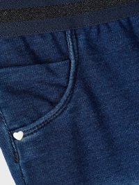 Denim ύφασμα σε σκούρο μπλε με μαύρη ελαστική μέση. Διαθέτει ένα μικρό μεταλλικό διακοσμητικό σε σχήμα καρδιάς στην τσέπη. Λείο υφή.