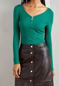 Haut vert côtelé à manches longues avec col en V et fermeture éclair frontale, associé à une jupe en cuir noire ornée de boutons-pression sur le devant.