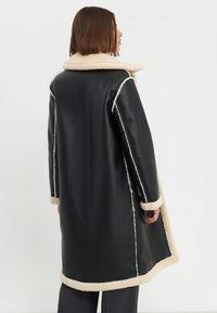 Cappotto lungo in finta pelle nera con colletto e polsini in finta pelliccia crema, caratterizzato da cuciture a contrasto e una silhouette rilassata.