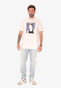 Homme avec des tatouages portant un t-shirt graphique blanc, un jean bleu clair et des baskets blanches, se tenant devant un fond uni.
