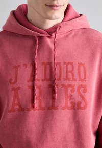 Różowa bluza z kapturem na sznurki, z napisem "J’ADORE ARIES" w ciemniejszym odcieniu. Miękka tekstura z casualowym stylem.