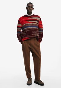Gestreifter Strickpullover in Rot, Maroon und Blau, kombiniert mit braunen, gefalteten Hosen und grau-braunen Schuhen. Verfügt über einen weiten Halsausschnitt und einen strukturierten Stoff.