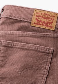 Pantalons en velours côtelé de couleur rose poussiéreux avec une surface texturée. Les caractéristiques comprennent une étiquette en cuir avec des détails en relief rouges et des poches arrière.