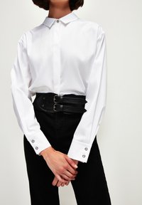 Camicia bianca a maniche lunghe con colletto piccolo, chiusa con un bottone argento. Pantaloni neri a vita alta con una cintura in pelle larga dotata di doppia fibbia.