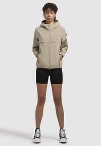 Šviesiai smėlio spalvos zip-up striukė su gobtuvu, elastiniais rankogaliais ir juosmeniu, su priekiniais kišenėmis ir minimalizmo dizainu, derinama su juodomis šortais ir sportbačiais.