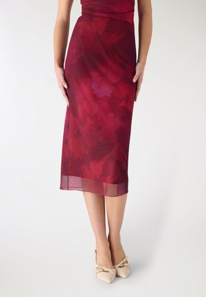 Donna che indossa una gonna trasparente rossa a fiori lunga fino al ginocchio e tacchi crema con punta appuntita e cinturini alla caviglia, in piedi su sfondo bianco.