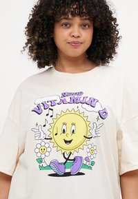 Cream-colored t-shirt featuring a cartoon sun graphic with a smiling face, surrounded by flowers and the text “NEED VITAMIN D” in purple.