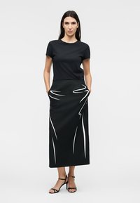Femme portant un haut noir à manches courtes et une jupe midi noire avec des lignes abstraites blanches, debout, les mains dans les poches de la jupe, talons noirs.