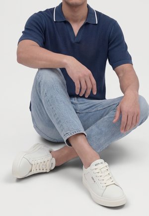 Homme assis en tailleur portant un polo bleu marine, un jean bleu clair et des baskets blanches Replay sur fond blanc.