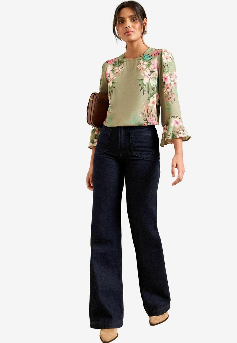 Olive green flower-patterned blouse with flared sleeves, paired with high-waisted dark denim jeans and beige heeled shoes. Brown handbag.