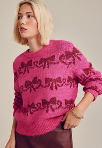 Rosa stickad tröja med strukturerad yta, med ett mönster av mörka rosetter. Ribbad krage och muddar, bärs över en vinröd kjol.