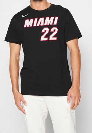 Mężczyzna ubrany w czarną koszulkę Nike Miami 22 i białe spodnie Nike z logo NBA, stojący na jednolitym tle.
