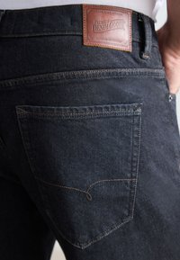 Tummat farkut, joissa on teksturoitu pinta, varustettu ruskealla nahkaleimalla, jossa lukee "Premium Denim Goods", sekä kontrastiväriset saumat takataskussa.