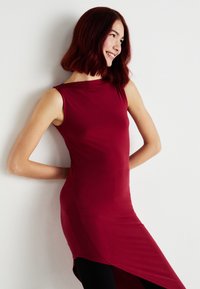 Vestido sem mangas Borgonha com um decote alto e bainha assimétrica, feito de um tecido suave. O modelo posa contra um fundo branco.