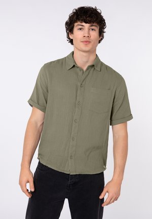 Jonge man met krullend haar die een olijfgroen, kort mouwig overhemd met knopen draagt en een zwarte broek, staand tegen een effen witte achtergrond.