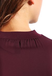 Haut bourgogne avec un décolleté modeste et un détail froncé au col arrière. Tissu lisse et léger avec une finition soignée.