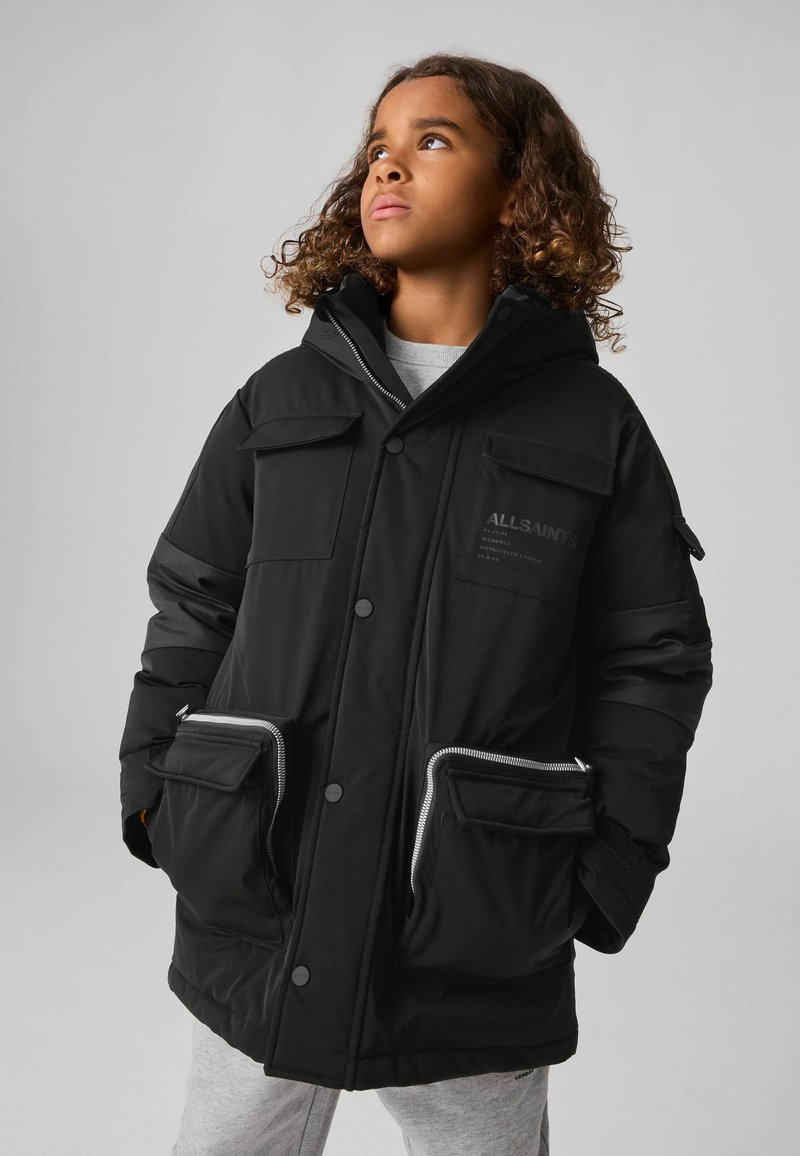 Chaqueta acolchada negra con capucha, que cuenta con múltiples bolsillos con cremallera y botón, una textura suave y un ajuste ligeramente holgado.