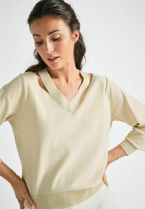 Woman wearing a beige V-neck sweater with cut-out shoulder details, looking down with hands resting on her hips.