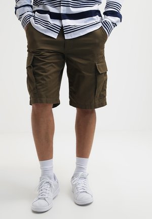 Person iført olivengrøn cargoshorts, hvide sokker, hvide sneakers og en blå- og hvide stribet langærmet skjorte med hænderne i lommerne.