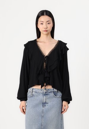 Black cropped blouse with ruffle detailing, deep V-neckline, and tie closure, featuring long sleeves with elastic cuffs and textured fabric.
