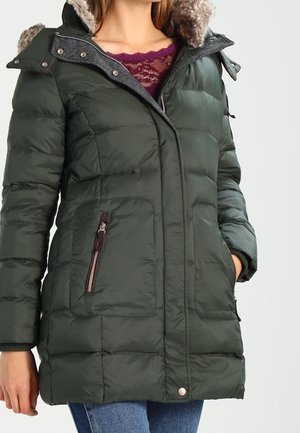 Femme portant un manteau d'hiver matelassé vert foncé avec capuche bordée de fourrure, poches zippées et un top en dentelle violet en dessous.