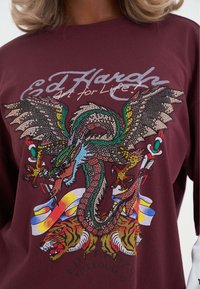 Bordowa koszulka z długim rękawem z żywym wzorem smoka i tygrysa, ozdobiona cekinami, umieszczona na tłem z nadrukiem.