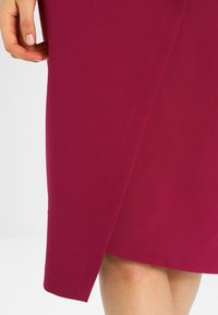 Enganliegendes magentafarbenes Kleid mit einem Schlitz an der Seite, aus glattem Stoff gefertigt. Die Nahaufnahme zeigt die Textur und die klaren Linien, die das elegante Design betonen.