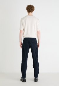 Beige T-Shirt mit kurzen Ärmeln und marineblauen Cargo-Hosen mit Taschen. Die Hose hat eine strukturierte Oberfläche und einen geraden Beinansatz. Schwarze Stiefel sind sichtbar.