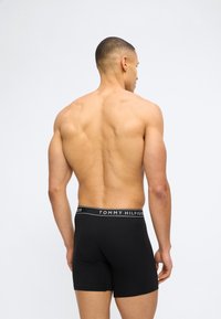 Μυώδης άνδρας στέκεται με την πλάτη στην κάμερα, φορώντας μαύρα μποξεράκια Tommy Hilfiger μπροστά σε λευκό, μονόχρωμο φόντο.