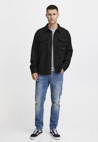 Black flannel shirt with two chest pockets, grey crew neck t-shirt underneath, light blue jeans, black sneakers with white accents.