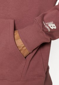 Main dans la poche d'un sweat New Balance rose fané avec poignets côtelés et logo blanc sur la manche.