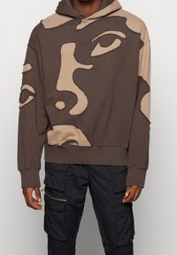 Felpa marrone e beige con motivi di volti astratti e pantaloni cargo neri con molte tasche e cerniere sulle gambe.