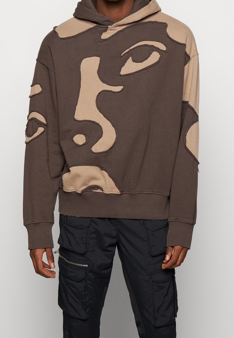 Felpa marrone e beige con motivi di volti astratti e pantaloni cargo neri con molte tasche e cerniere sulle gambe.