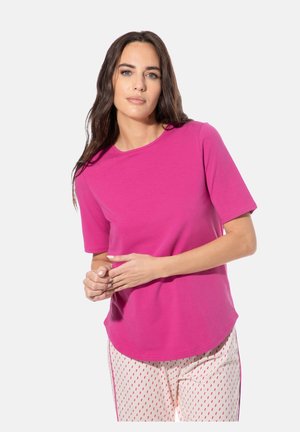 Frau mit langen dunklen Haaren, die ein hellrosa kurzärmeliges Shirt und helle Hosen mit kleinen roten Mustern vor einem einfarbigen Hintergrund trägt.