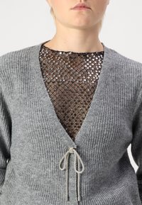 Hallux ribbes koot, millel on sügava V-kaelusega, metallist nööriga aksessuaar ja sädelevast seelikust aluskihist.