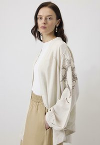 Cardigan bej cu deschidere frontală, decorated with accente din dantelă cu model pe mâneci, dintr-un material ușor și cu mansete pliate. Asortat cu un top crem și pantaloni bej.