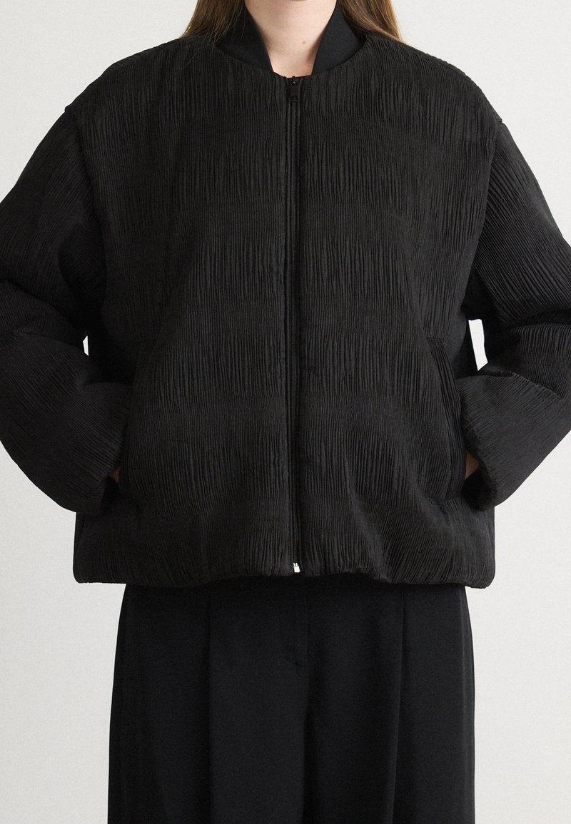 Veste bomber noire texturée avec fermeture éclair avant portée par une personne les mains dans les poches, associée à un pantalon noir sur fond uni.