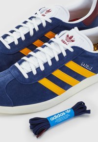 Blå mocka Adidas Gazelle-sneakers med gula ränder, vita snören och texturerad gummisula. Inkluderar ett mörkblått skosnöre.
