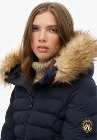 Navy padded winter jacket with faux fur hood, zippered front, and ribbed high-neck sweater underneath. Features logo patch on sleeve.