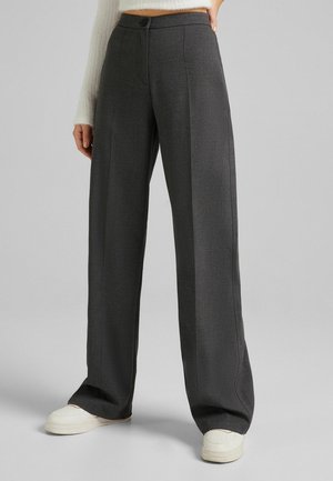 Personne portant un pantalon gris foncé à jambes larges et des baskets blanches, une main posée sur la hanche, debout devant un fond uni.