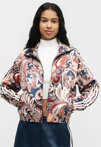 Jeune femme aux longs cheveux foncés porte un crop top blanc et une veste zippée colorée à motif cachemire avec un pantalon bleu marine.