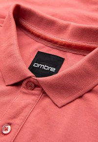 Korallfärgad polo-tröja med en strukturerad tyg, med en svart märkesetikett, två knappar och en traditionell kragsdesign.