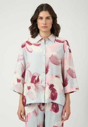 Kvinne iført en løs, knappet skjorte og matchende bukser med store rosa og burgunder blomster på lys blå bakgrunn.