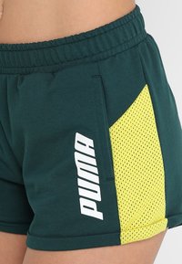 Pantalones cortos azul verdoso con cintura elástica, panel lateral de malla amarilla y logotipo vertical blanco de PUMA en la cadera y parte superior del muslo de una persona.
