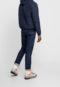 Navy puffer jacket with hood, worn over dark blue jeans. Tan and orange boots with laces and a textured sole. Faded cuff at jeans' hem.