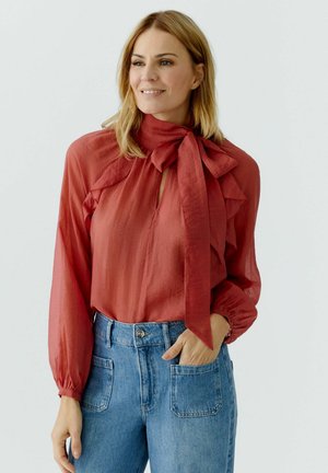 Woman wearing a rust-red long-sleeve blouse with a large bow at collar, tucked into high-waisted blue jeans with front patch pockets.